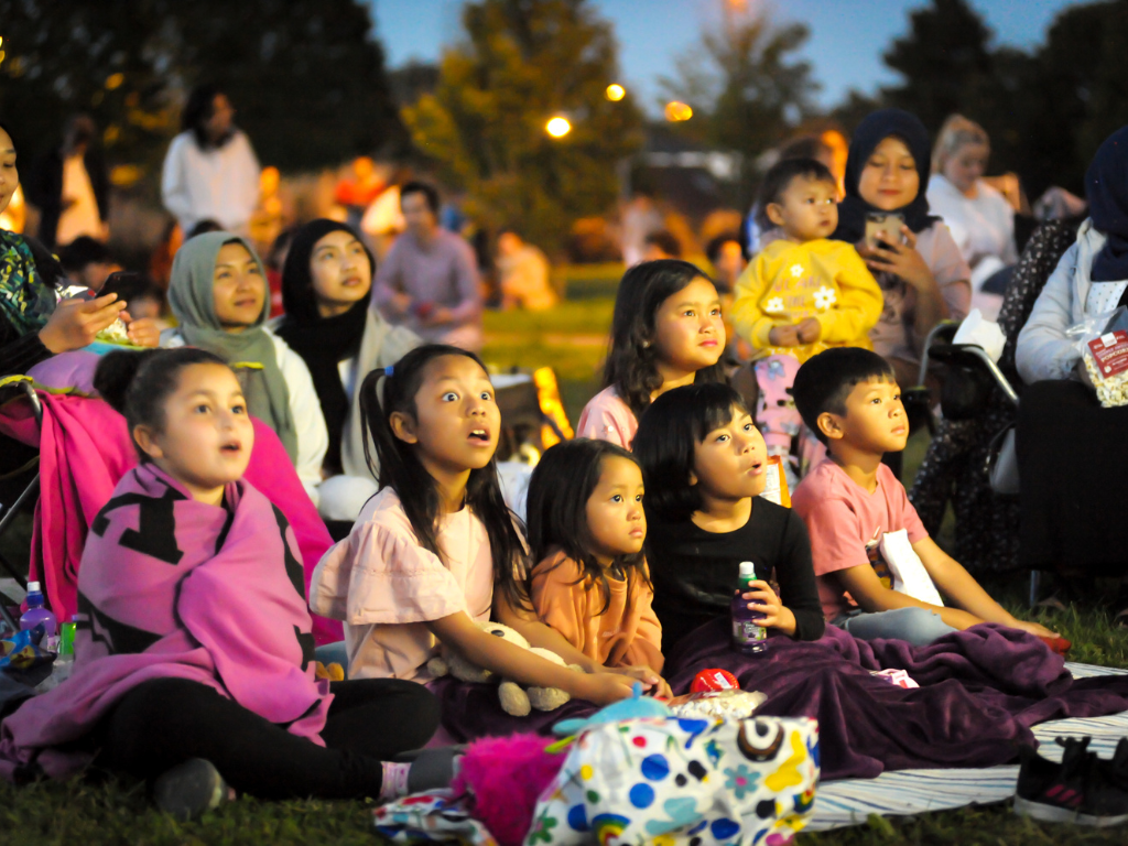A family at an outdoor film screening sit with awestruck children.
