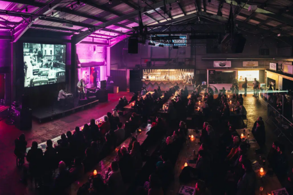 wide shot of an outdoor screening space with a packed audience watching a black and white film. There is striking purple lighting around the screen adding to the sense of atmosphere.