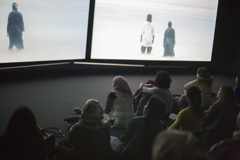 Audience sits on a floor viewing a two screen video installation