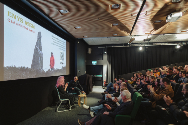 Photo of cinema from the side aisle showing a packed audience and two speakers sitting on stage. The cinema screen has a holding slide for Enys Men
