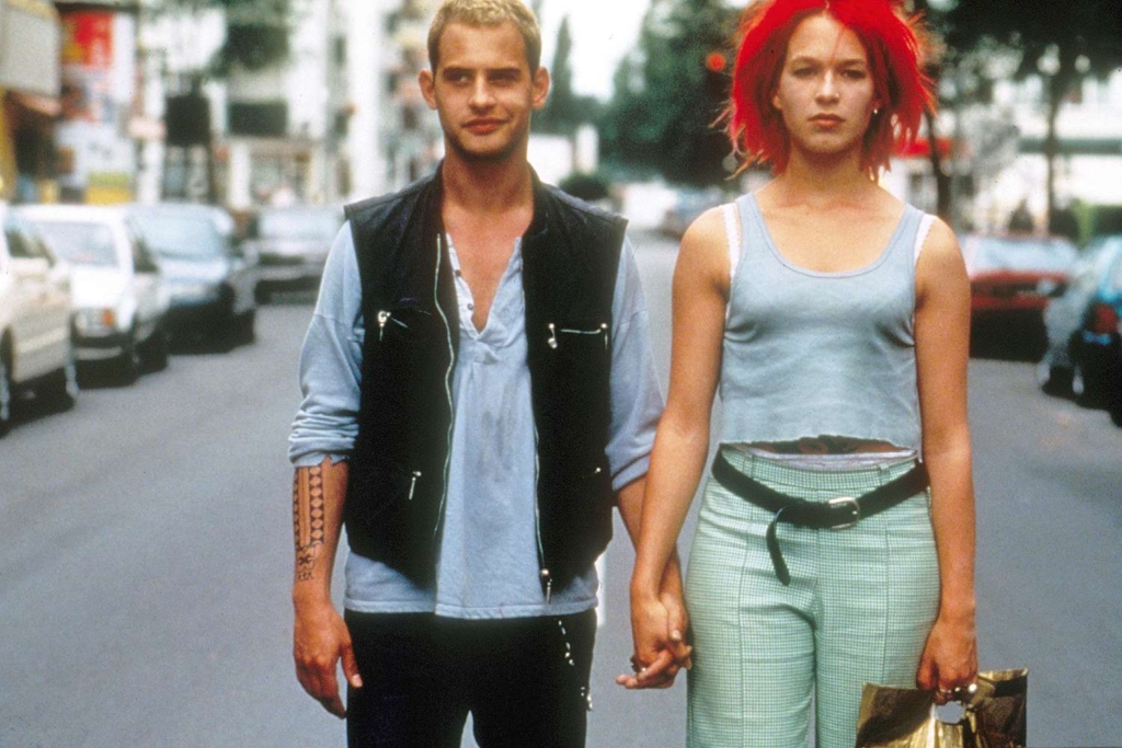 A man in a black waistcoat holds hand with a woman in with bright red hair on a street. She holds a plastic bag.