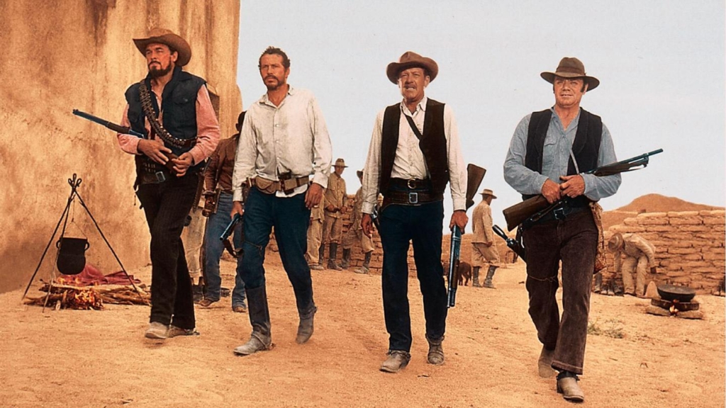 Four cowboys walk through a settlement holding rifles.