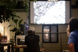 An old projector is playing a film on a screen. People stand beside the projector, watching the images.