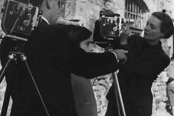 A black and white image of a woman adjusting a camera. A man and a boy stand beside her