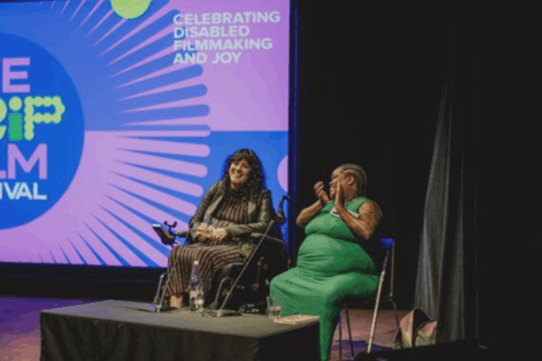 Kyla Harris and Tara Brown in conversation at We Crip Film Festival 2025. They sit on chairs in front of a purple and blue screen featuring the We Crip Film Festival logo and the text: Celebrating Disabled Filmmaking and Joy.