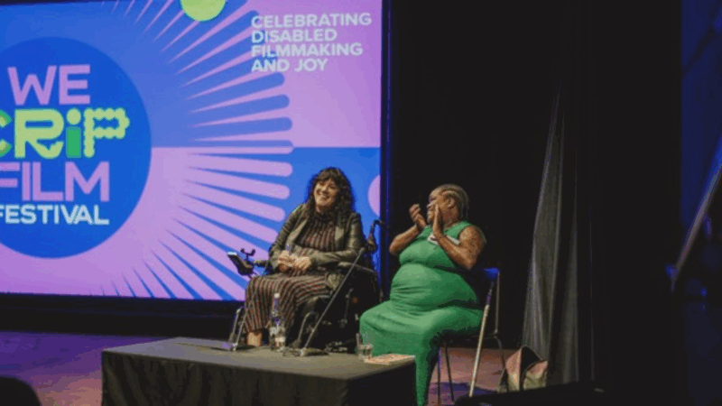 Kyla Harris and Tara Brown in conversation at We Crip Film Festival 2025. They sit on chairs in front of a purple and blue screen featuring the We Crip Film Festival logo and the text: Celebrating Disabled Filmmaking and Joy.