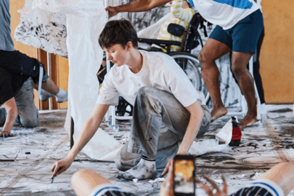 A young person in paint-covered clothes is using a brush and painting on a canvas on the floor. Other people are also painting around them.