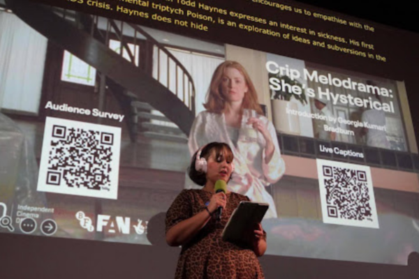 Image description: Close-up from Crip Melodrama’s launch event at the Barbican. Georgia Bradburn is wearing headphones and holding a microphone and a tablet. She’s speaking into the microphone and standing in front of a cinema screen. The screen’s holding slide features an image from the film Safe, showing Carol (played by Julianne Moore) standing in a modern kitchen, holding a glass of milk, and wearing a satin robe. There are two QR codes on each slide: one for live captions and one for the survey. There’s large white text that reads “Crip Melodrama: She’s Hysterical”. At the bottom of the slide are the logos for BFI FAN and the ICO. At the top of the screen is the live-caption overlay for Georgia’s introduction.