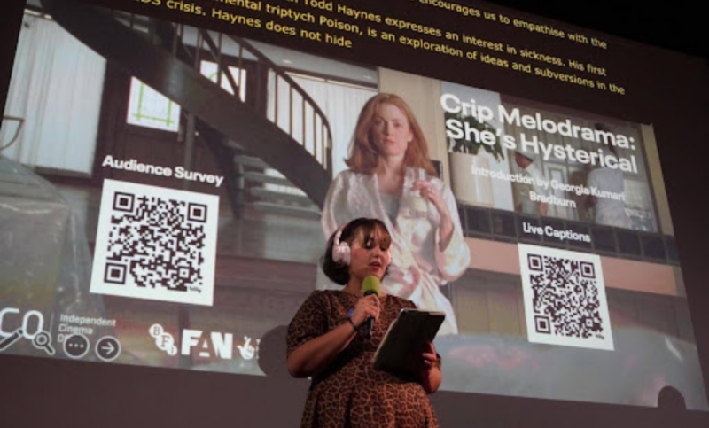 Image description: Close-up from Crip Melodrama’s launch event at the Barbican. Georgia Bradburn is wearing headphones and holding a microphone and a tablet. She’s speaking into the microphone and standing in front of a cinema screen. The screen’s holding slide features an image from the film Safe, showing Carol (played by Julianne Moore) standing in a modern kitchen, holding a glass of milk, and wearing a satin robe. There are two QR codes on each slide: one for live captions and one for the survey. There’s large white text that reads “Crip Melodrama: She’s Hysterical”. At the bottom of the slide are the logos for BFI FAN and the ICO. At the top of the screen is the live-caption overlay for Georgia’s introduction.