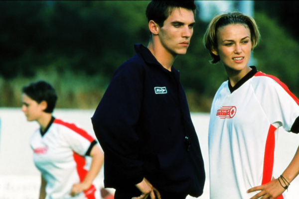 A still from Bend It Like Beckham: Two young people stand on a soccer field; one wears a dark jacket, the other a white jersey. Another player is blurred in the background.
