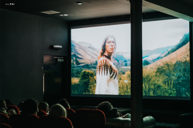 Photo of an audience watching a cinema screen with a woman standing in the mountains on screen.